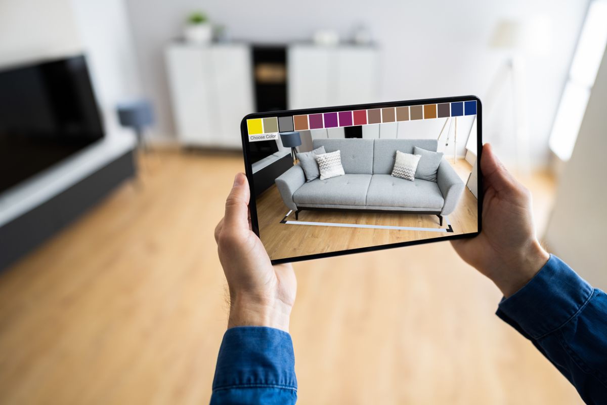 Ruky držiace tablet s aplikáciou rozšírenej reality na vizualizáciu sivej pohovky a farieb v izbe.