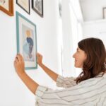 Žena v bielej košeli vešia obraz s abstraktnými modrými a oranžovými tvarmi na bielu stenu v interiéri.