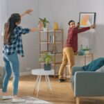 Muž s kockovanou košeľou vešia obraz na bielu stenu, zatiaľ čo mu žena radí v obývačke so sivými kreslami.