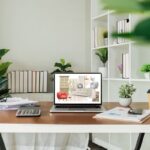 Pracovný stôl s notebookom zobrazujúcim inšpiračnú koláž interiérových prvkov a rastlín na policiach.