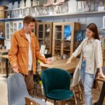 Pár v obchode s nábytkom si prezerá tmavozelené a sivé čalúnené stoličky pri drevenom jedálenskom stole.