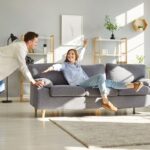 Mladý pár presúva v obývačke sivú pohovku na biely koberec s dreveným regálom a svetlom.