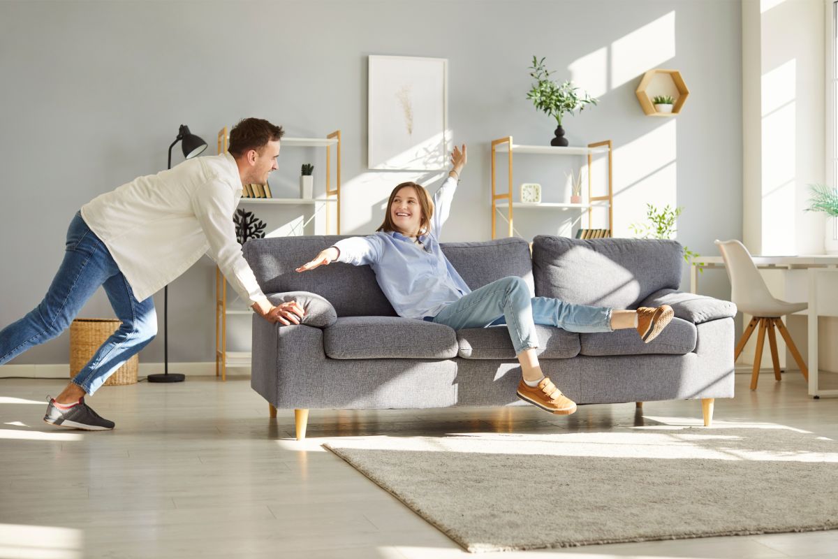 Mladý pár presúva v obývačke sivú pohovku na biely koberec s dreveným regálom a svetlom.