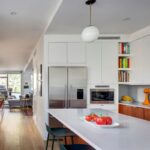 Moderná biela kuchyňa so vstavanou chladničkou z nehrdzavejúcej ocele a výhľadom do otvorenej obývacej izby.