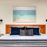 Spálňa s dreveným čelom postele, veľkým obrazom tyrkysového mora a čiernymi nástennými lampami.