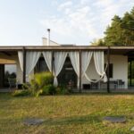 Moderný prízemný dom so sedlovou strechou a krytou terasou so závesmi pri upravenom trávniku.