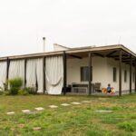 Moderný prízemný dom so sedlovou strechou a krytou terasou so závesmi pri upravenom trávniku.