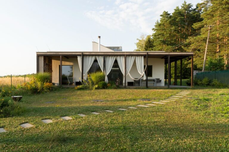 Moderný prízemný dom so sedlovou strechou a krytou terasou so závesmi pri upravenom trávniku.