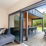 Pohľad z interiéru na terasu s drevenou palubovkou, tmavosivou vonkajšou pohovkou a pergolou s dreveným obkladom a kovovou strechou.