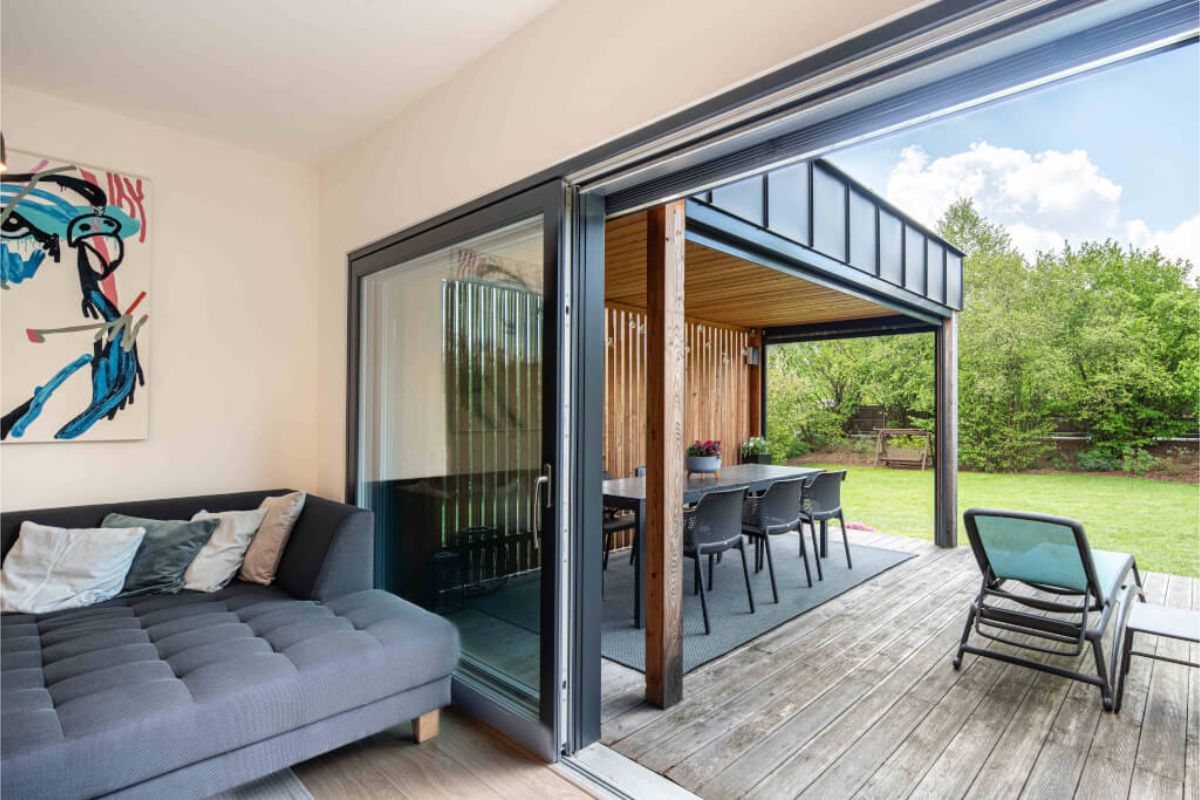 Pohľad z interiéru na terasu s drevenou palubovkou, tmavosivou vonkajšou pohovkou a pergolou s dreveným obkladom a kovovou strechou.