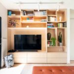 Interiérová stena s veľkým vstavaným TV nábytkom zo svetlej drevenej preglejky a hnedou koženou taburetkou pred posuvnými dverami na terasu.