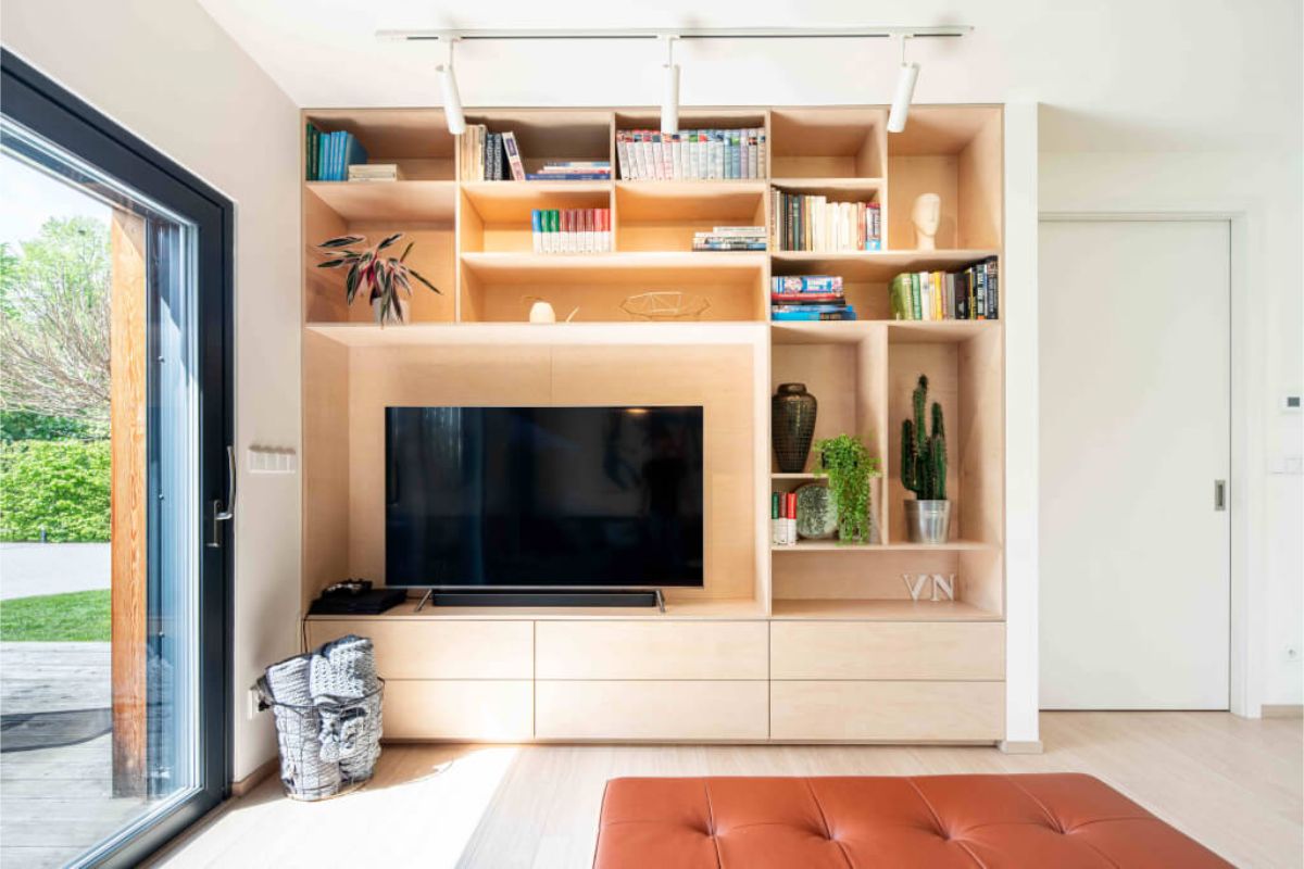 Interiérová stena s veľkým vstavaným TV nábytkom zo svetlej drevenej preglejky a hnedou koženou taburetkou pred posuvnými dverami na terasu.