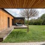 Vonkajší pohľad na rohovú drevenú terasu s ležadlami a mladým stromčekom, obklopenú sviežim zeleným trávnikom a dreveným obkladom domu.