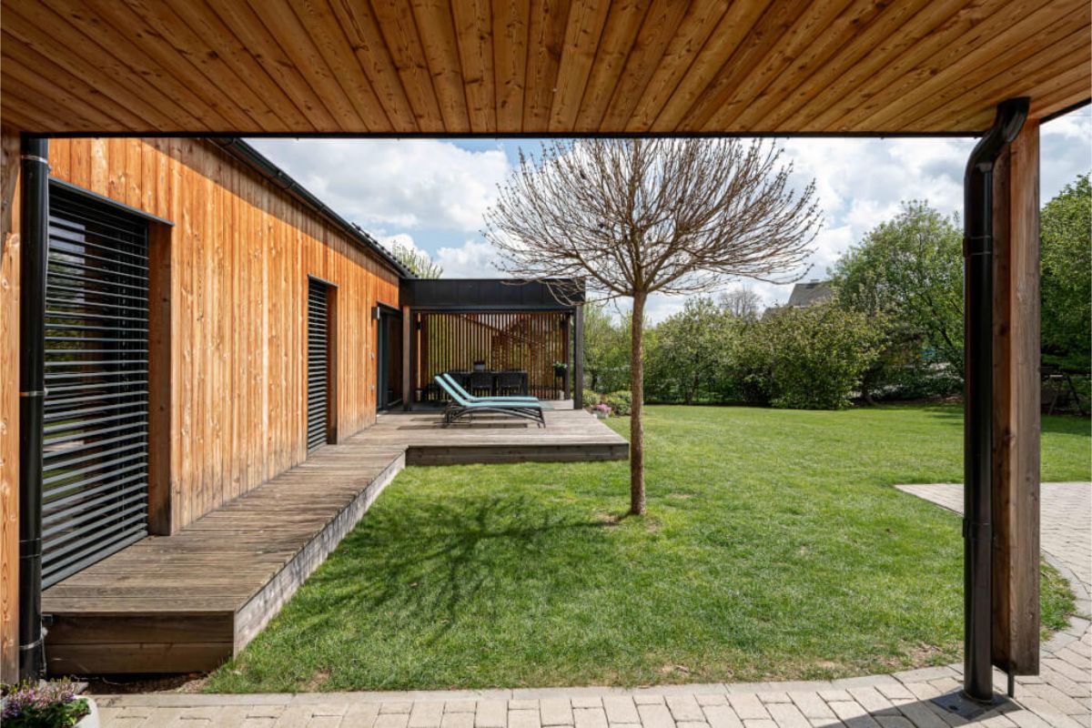 Vonkajší pohľad na rohovú drevenú terasu s ležadlami a mladým stromčekom, obklopenú sviežim zeleným trávnikom a dreveným obkladom domu.