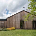 Moderný rodinný dom s dreveným obkladom, tmavou šikmou plechovou strechou a nenápadnými čiernymi oknami, zasadený v zelenej záhrade.