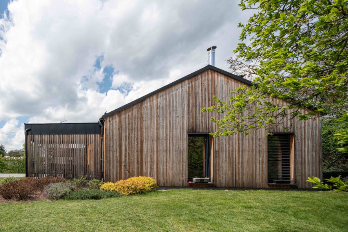Moderný rodinný dom s dreveným obkladom, tmavou šikmou plechovou strechou a nenápadnými čiernymi oknami, zasadený v zelenej záhrade.