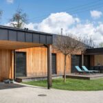 Exteriér domu s čiastočným dreveným obkladom, svetlou betónovou dlažbou pred vchodom a terasou so záhradnou pergolou a ležadlami.