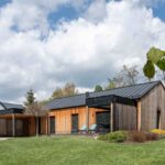 Nízky moderný dom so zvislým dreveným obkladom, čiernou strechou a prístreškom pre auto, obklopený trávnatým pozemkom.