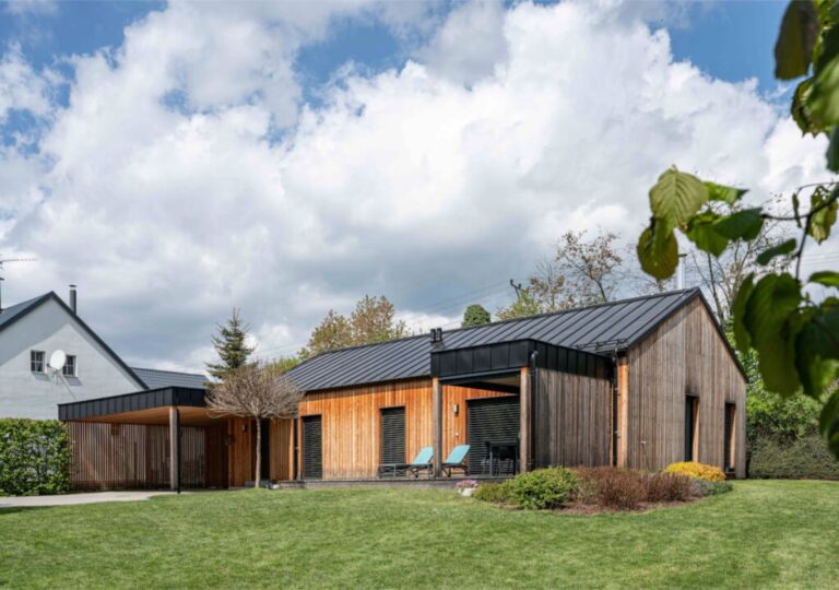 Nízky moderný dom so zvislým dreveným obkladom, čiernou strechou a prístreškom pre auto, obklopený trávnatým pozemkom.