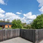 Predná časť pozemku s tmavou drevenou posuvnou bránou a plotom s dláždenou prístupovou cestou, v pozadí viditeľný moderný drevený dom.