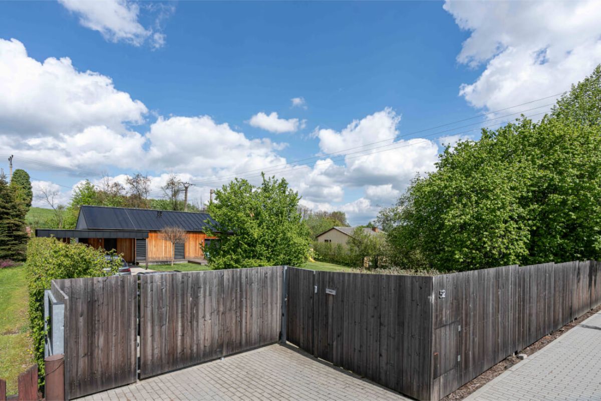 Predná časť pozemku s tmavou drevenou posuvnou bránou a plotom s dláždenou prístupovou cestou, v pozadí viditeľný moderný drevený dom.