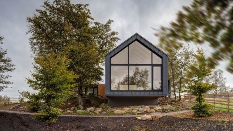 Moderný chalet so sivou fasádou a preskleným štítom v prírodnom prostredí.