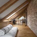 Podkrovný priestor s drevenou podlahou, priznanými trámami, kamenným obkladom a presklenou stenou do kancelárie.