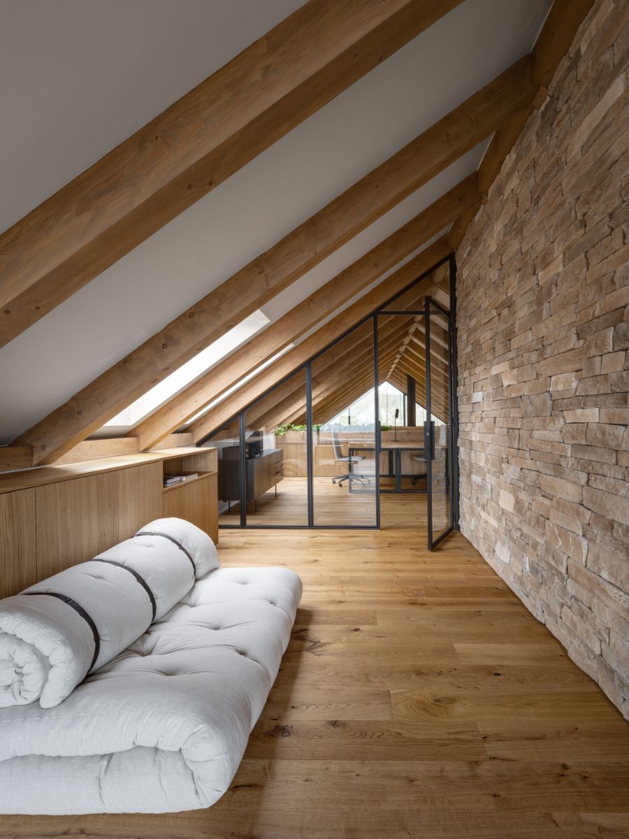 Podkrovný priestor s drevenou podlahou, priznanými trámami, kamenným obkladom a presklenou stenou do kancelárie.