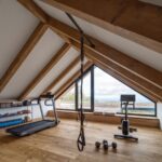 Domáca posilňovňa v podkroví s drevenou podlahou, bežeckým pásom, trámami a oknom s výhľadom na krajinu.