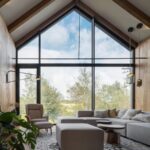 Obývacia izba so šikmým stropom, drevenými trámami, veľkou presklenou stenou svetlosivou sedačkou.