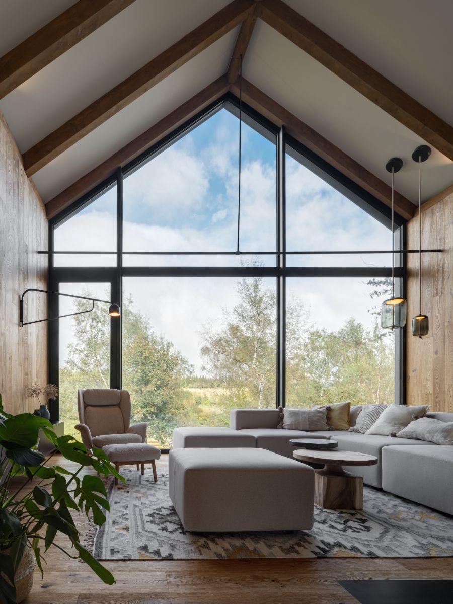 Obývacia izba so šikmým stropom, drevenými trámami, veľkou presklenou stenou svetlosivou sedačkou.