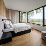 Spálňa s drevenou podlahou a obkladom, nízkou posteľou a veľkým rohovým oknom s prírodným výhľadom.