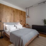 Moderná spálňa s drevenu stenou, sivou posteľou, čiernymi nočnými stolíkmi a bielou stenou.