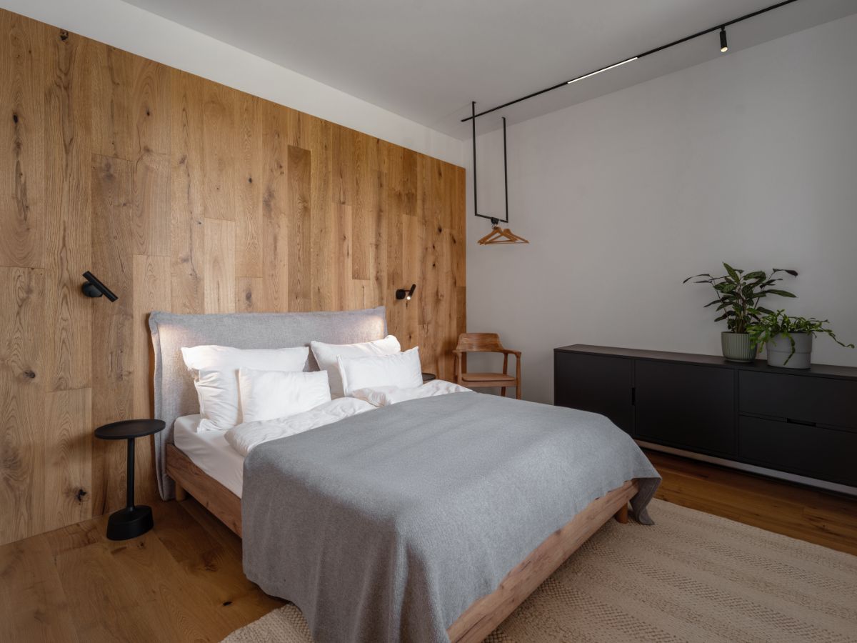 Moderná spálňa s drevenu stenou, sivou posteľou, čiernymi nočnými stolíkmi a bielou stenou.