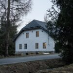 Uličný pohľad na prednú fasádu vidieckej chaty s bielym štukom, šedými okennými rámami a kamennou podmurovkou.