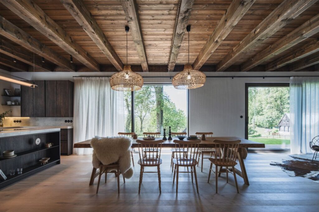 Masívny drevený stôl s dizajnovými stoličkami v otvorenej jedálni s trámovým stropom, prútenými lustrami a veľkoformátovým oknom do záhrady.