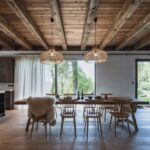 Masívny drevený stôl s dizajnovými stoličkami v otvorenej jedálni s trámovým stropom, prútenými lustrami a veľkoformátovým oknom do záhrady.