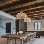 Moderná rustikálna jedáleň s masívnym dreveným stolom, tmavou kuchynskou linkou v pozadí a priznaným trámovým stropom s prútenými svietidlami.