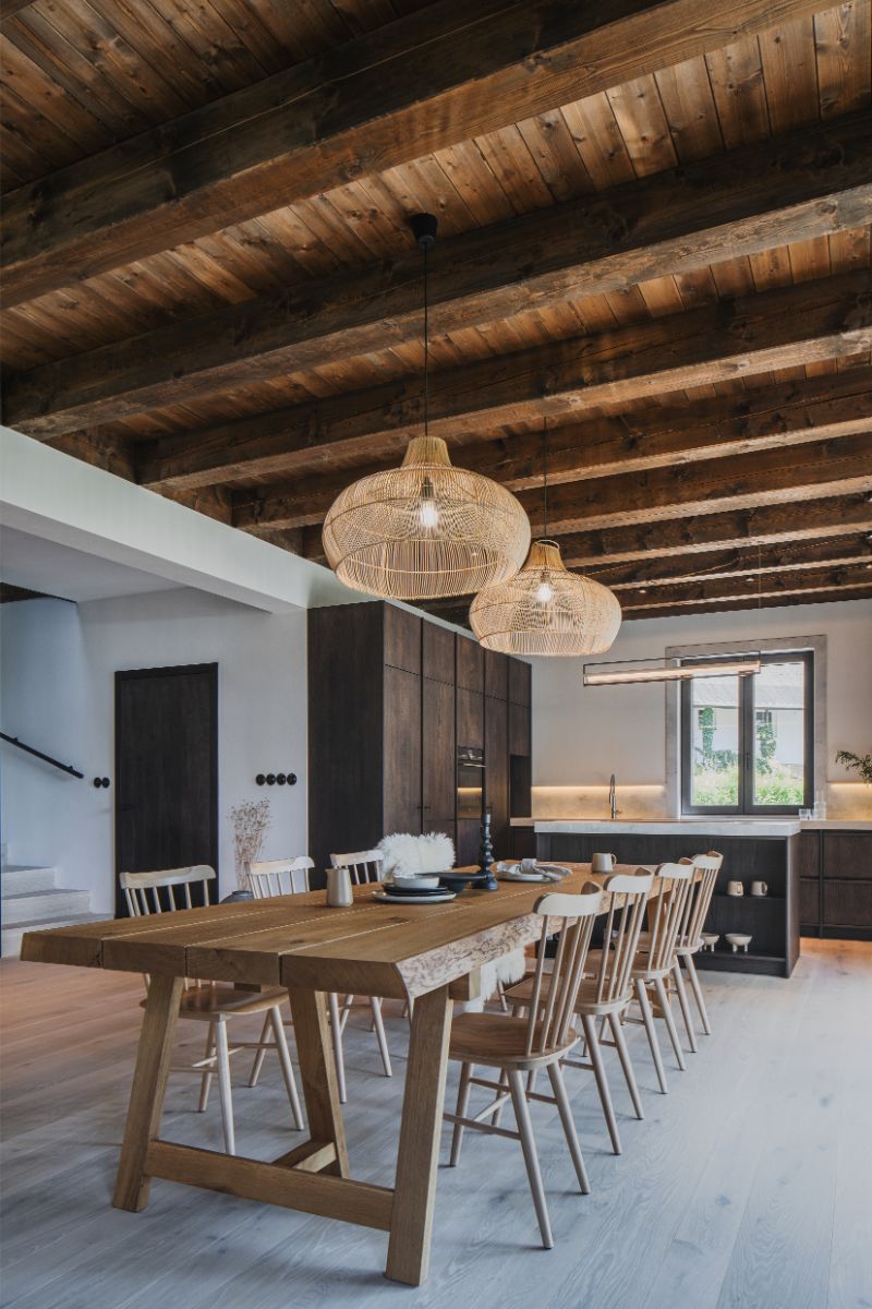 Moderná rustikálna jedáleň s masívnym dreveným stolom, tmavou kuchynskou linkou v pozadí a priznaným trámovým stropom s prútenými svietidlami.