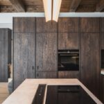 Moderná kuchynská zostava z tmavého štruktúrovaného dreva so zabudovanými spotrebičmi a svetlou kamennou pracovnou doskou na ostrove.