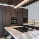 Kuchynský ostrov so svetlou kamennou doskou, indukčnou varnou doskou a tmavým dreveným nábytkom v modernom horskom interiéri.