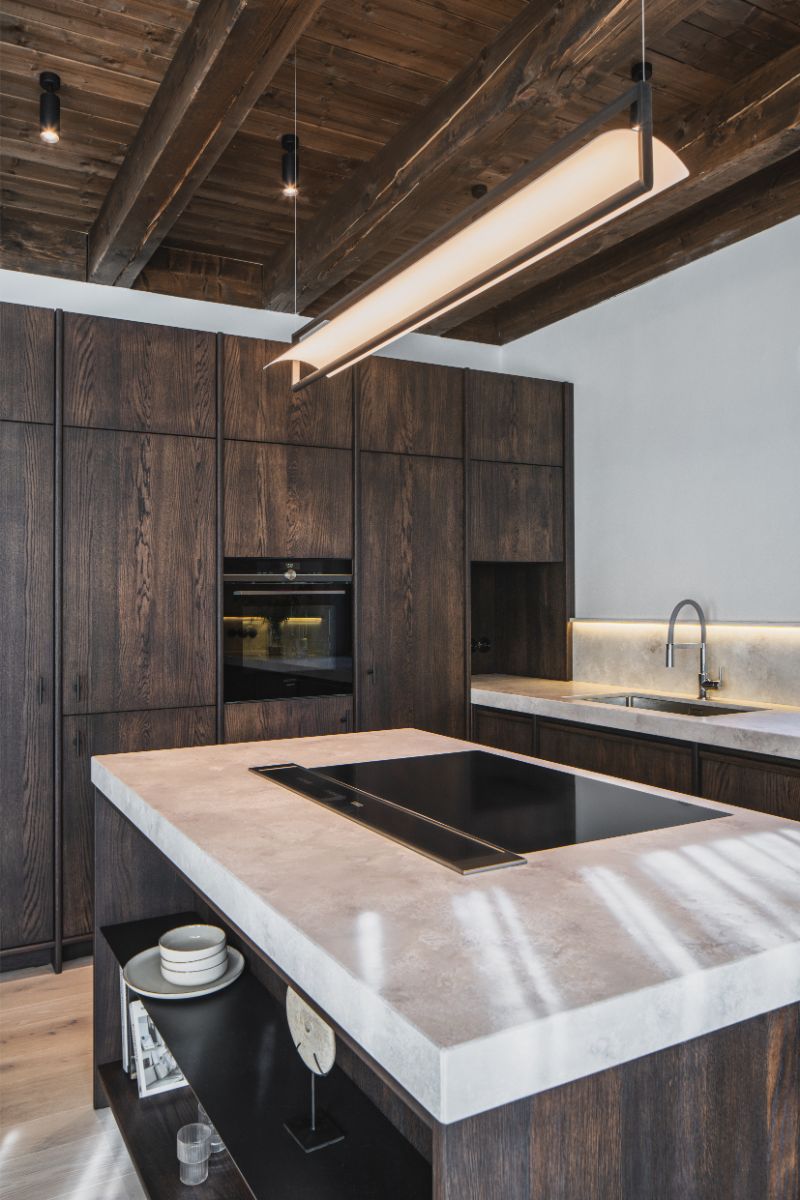 Kuchynský ostrov so svetlou kamennou doskou, indukčnou varnou doskou a tmavým dreveným nábytkom v modernom horskom interiéri.