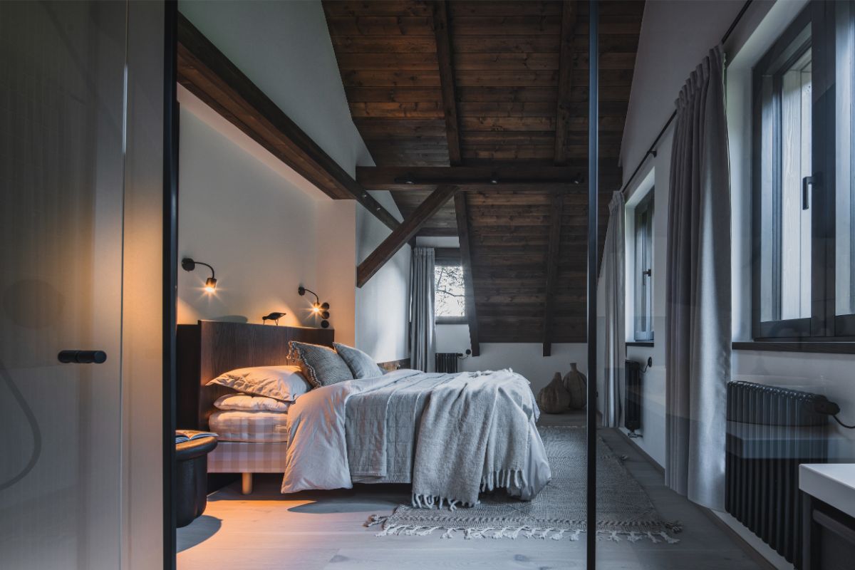 Podkrovná spálňa s masívnym trámovým stropom, posteľou v neutrálnych tónoch, dizajnovými lampami a čiastočne presklenou stenou do kúpeľne