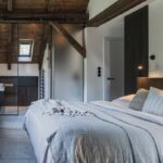 Moderná spálňa v podkroví s dreveným obkladom stropu, svetlou posteľnou bielizňou a priehľadom cez sklenenú priečku do kúpeľne s umývadlom.