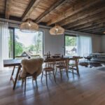 Otvorený spoločenský priestor s veľkým dreveným stolom, prútenými lustrami, trámovým stropom a výhľadom cez veľkoformátové okná do prírody.