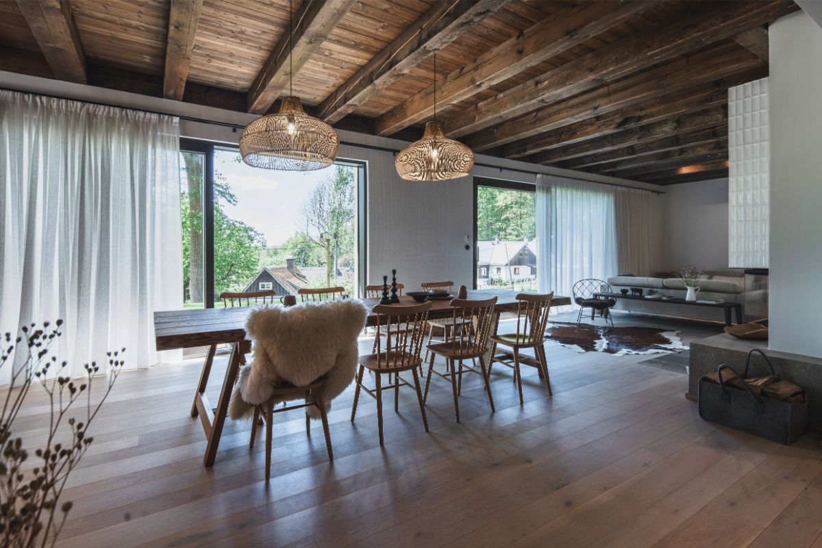 Otvorený spoločenský priestor s veľkým dreveným stolom, prútenými lustrami, trámovým stropom a výhľadom cez veľkoformátové okná do prírody.