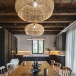 Kuchyňa s tmavou linkou prepojená s jedálňou s masívnym stolom, prútenými svietidlami a drevenými trámami na strope.