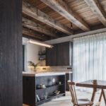 Kuchynský ostrov z tmavého dreva so svetlou doskou v interiéri s trámovým stropom, lineárnym svietidlom a bielymi ľanovými závesmi.