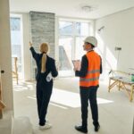 Mladá žena s papierom pod pazuchou a pracovník v reflexnej veste a prilbe v rekonštruovanej miestnosti.