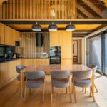 Moderná kuchyňa a jedáleň v chate s drevenými obkladmi, trámami a šedými stoličkami pri lesklom drevenom stole.